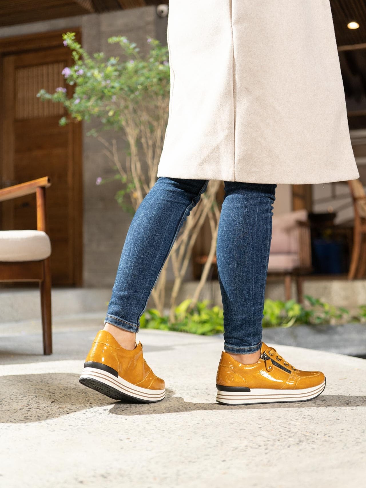 Gelbe Sneaker aus Kunstlack mit Reißverschluss und Schnürung und Wechselfußbett. Passend zu den Schuhen trägt die Frau einen hellen Mantel und eine enge Jeanshose.