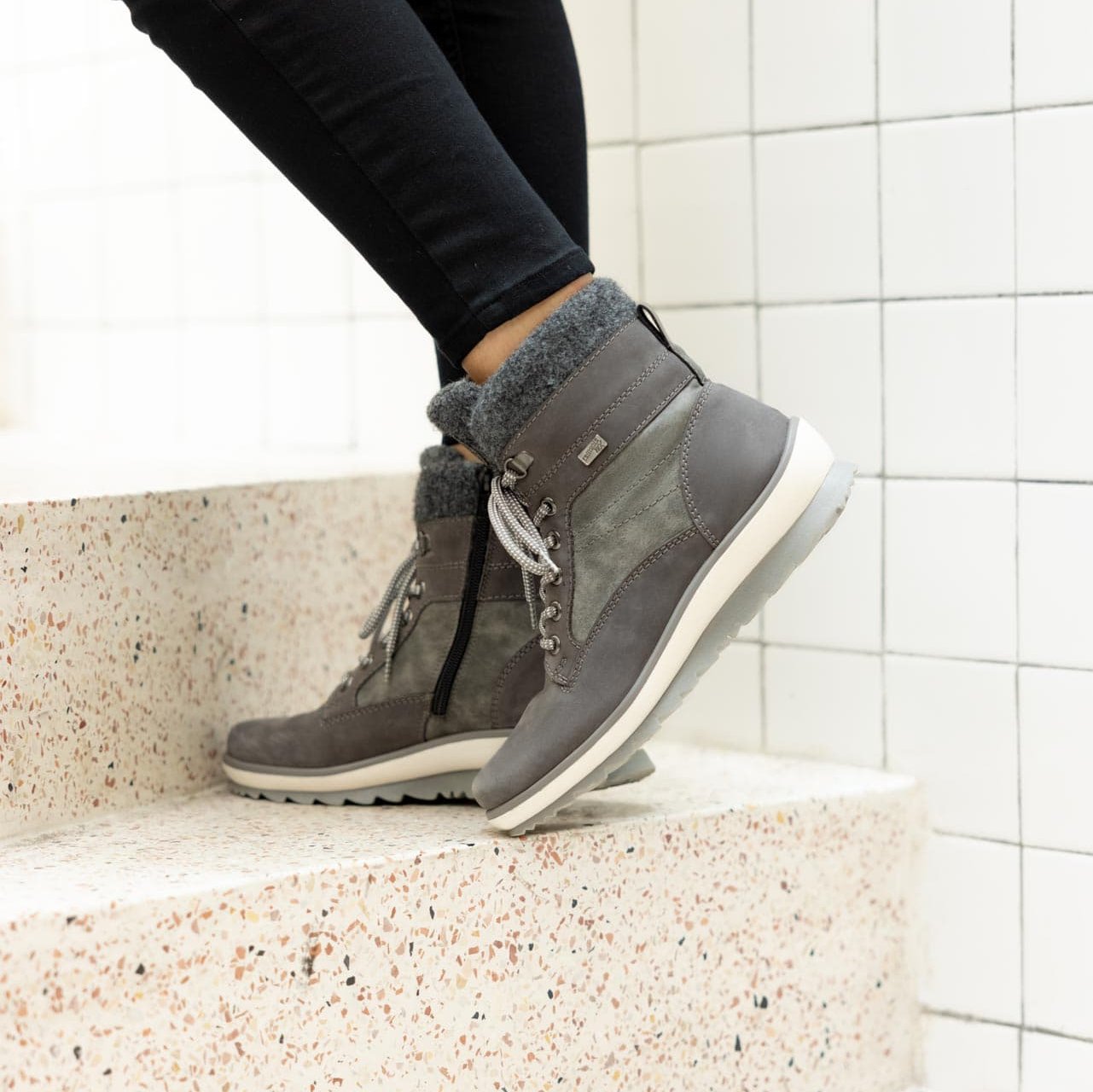 Graue Stiefel warm gefüttert aus Rauhleder mit Reißverschluss und Schnürung, wasserabweisendem Remonte TEX und Wechselfußbett. Passend zu den Schuhen trägt die Frau im Treppenhaus eine schwarze Hose.