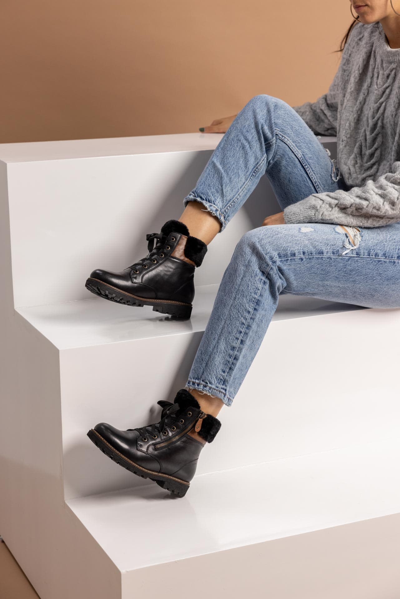 Schwarze Stiefeletten warm gefüttert aus Glattleder mit Reißverschluss und Schnürung und Wechselfußbett. Passend zu den Schuhen trägt die Frau auf der Treppe einen grauen Strickpullover und eine helle Mom-Jeans.